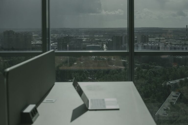 Laptop on office desk with a city skyline through a large window, offering a modern workspace view.