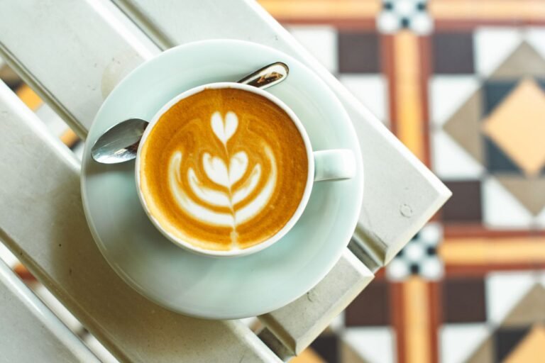 A top-down view of a latte with artistic foam in a white cup placed on a patterned tile surface.
